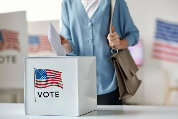 stock image Young female citizen of United States of America putting paper into ballot box while standing by desk in polling place and making her choice