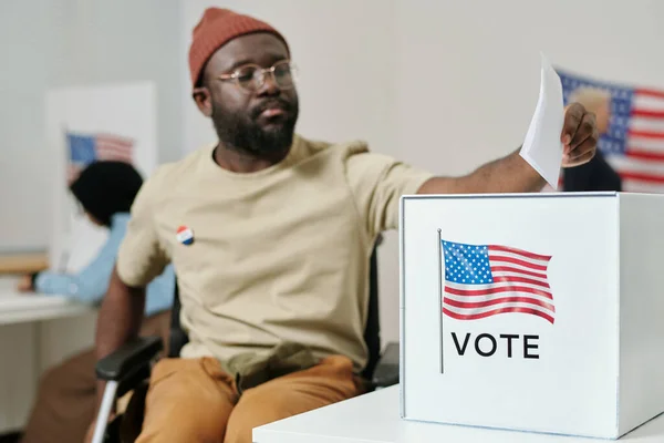Box for ballot papers on desk and young African American man with disability sitting in wheelchair and making his choice