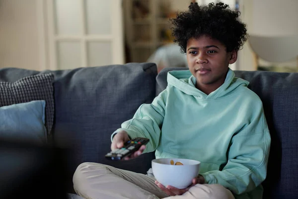 有一碗薯片的可爱的非裔美国男孩坐在沙发上选择电视频道 并按下遥控器上的按钮 — 图库照片