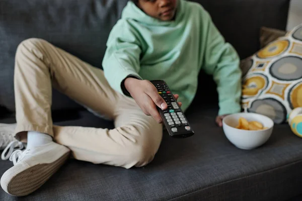 軍隊ショットの小さい男の子でパーカーとパンツ選択テレビチャンネルながら座っています上のソファでリビングルームとプレスボタンオンリモートコントロール — ストック写真