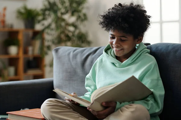 Usmívající Afroameričan Školák Casualwear Čtení Knihy Zatímco Sedí Gauči Obývacím — Stock fotografie
