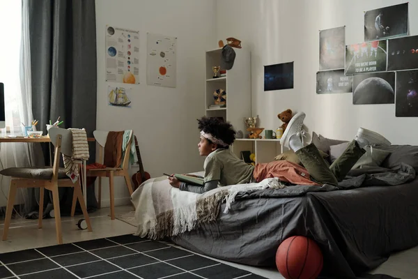 身穿便服的非洲裔美国男孩在舒适的双人床上放松下来 一边看电脑屏幕一边看录像 — 图库照片