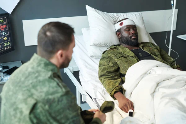 身着迷彩服的年轻黑人伤员躺在医院病房的床上 而他的朋友则坐在他面前参观 — 图库照片