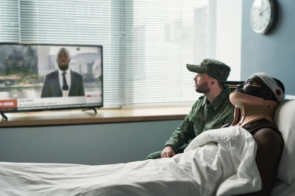 病院病棟の画面の前に座っている間にテレビでニュースを見て軍の制服を着た負傷した男と彼の仲間の側面図 — ストック写真