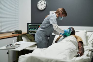 Koruyucu maskeli, eldivenli ve üniformalı hemşire hasta yatağında yatarken solunum maskesi takıyor.