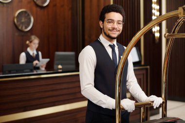 Genç, zarif bellboy üniformalı müşterilerin bagajlarını taşırken otel odasında hareket ediyor ve kameraya bakıyor.