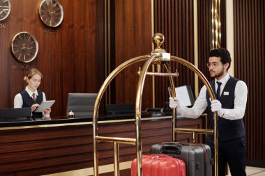 Genç bellboy, kadın yöneticiyle birlikte dijital tablet kullanarak resepsiyon tezgahında yürürken çantalarla el arabası itiyor.