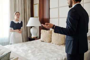 Lüks otelin müdürü, oda hizmetçisi üniformalı hizmetçiye talimat veriyor. Konuklar için çift kişilik yatağın diğer tarafında hazır bekliyor.