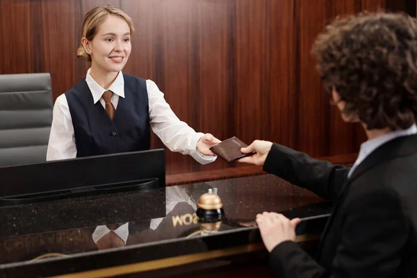 Happy young hotel staff looking at client passing her document over counter while standing by workplace and registrating new guests