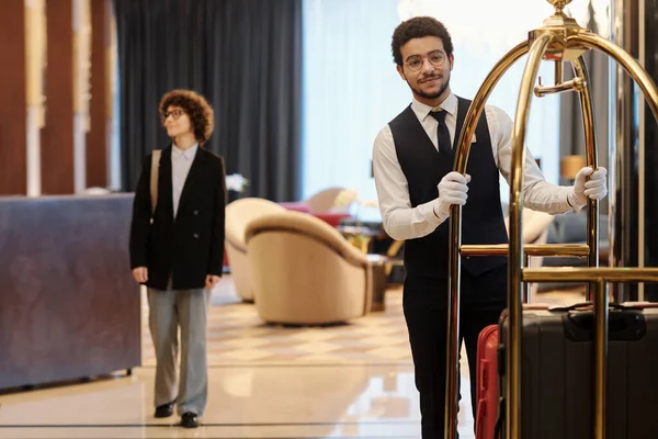 若いベルボーイはスーツケースを背景に女性旅行者と一緒にラウンジに沿って移動しながら ホテルのゲストの多くのスーツケースとカートを押す — ストック写真