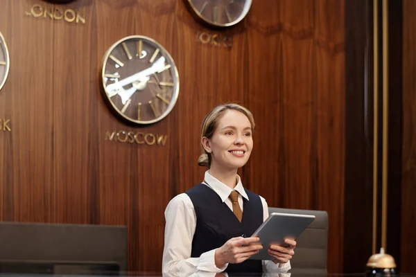 stock image Young smiling blond female receptionist of luxurious hotel holding tablet while standing by counter and waiting for new guests and travelers
