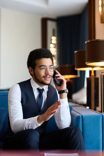 Young elegant male entrepreneur arranging appointment with business partner on mobile phone in cafe or lounge of luxurious hotel