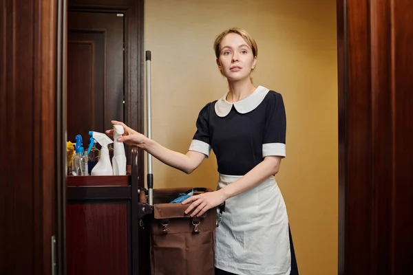 Junge Hübsche Zimmermädchen Uniform Nehmen Flasche Mit Sprayer Aus Einkaufswagen — Stockfoto