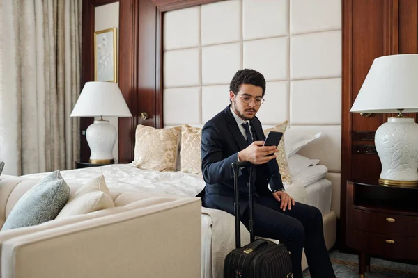 到着後ホテルの部屋でダブルベッドに座っている間 スマートフォンで正式に読書メッセージの若い重大なビジネスマン — ストック写真