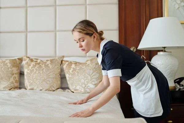 新しい旅行者のための豪華な5つ星ホテルの部屋を準備しながら ダブルベッドにきれいなベッド服を置く若いメイドのサイドビュー — ストック写真