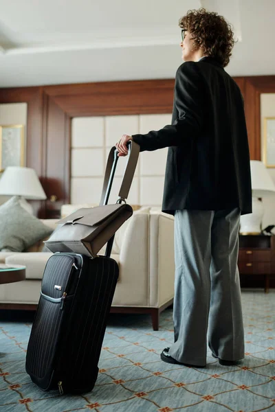 若いですブルネットビジネスマン女性でグレーパンツと黒ブレザースタンディングでホテルの部屋と保持スーツケースとともに彼女の手荷物によってハンドル — ストック写真