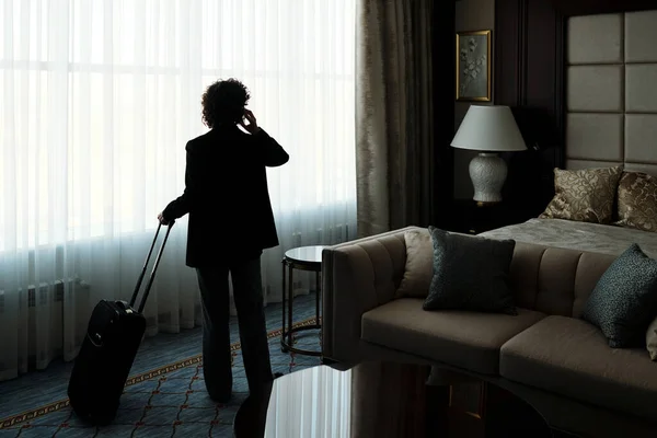 ホテルの部屋に白いシフォンカーテンで覆われた大きな窓の前に立っている間 スマートフォンで話しているビジネスマンの背面ビュー — ストック写真