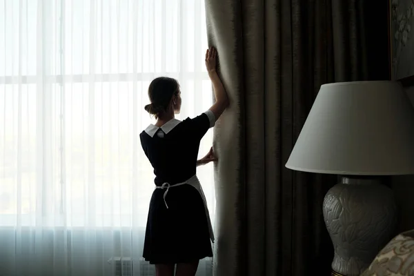 朝の大きな窓の前に立っている間 ホテルの部屋で重い夜のカーテンを開く制服を着た若い侍女のバックビュー — ストック写真