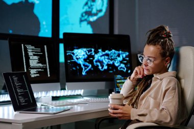Gözlüklü ciddi bir genç programcı bilgisayardaki kodlanmış verilere bakıyor ve iş yerinde karanlık ofiste otururken kahve içiyor.