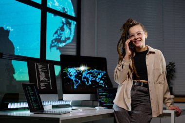 Akıllı telefonlu genç bir kadın gecenin geç saatlerinde karanlık ofiste bilişim destek şirketinin iş arkadaşı ya da müşterisiyle konuşurken gülüyor.