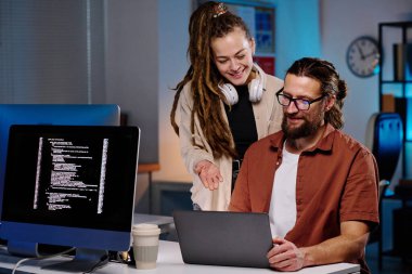 Genç, gülümseyen bir kadın bilgisayarın ekranını gösterirken aynı zamanda karanlık ofiste iş yerinde toplanan erkek arkadaşa verileri açıklıyor.