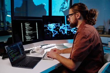 Genç ve ciddi bir erkek programcı karanlık ofiste çalışırken bilgisayar ekranında şifreli verilere bakıyor.