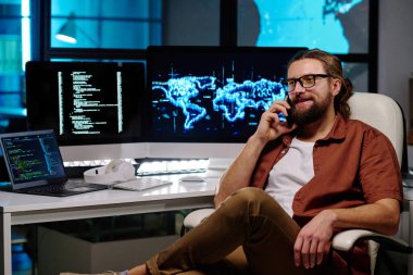 Genç gülümseyen bilişim yöneticisi iş yerinde ekranlarda şifreli verileri olan bilgisayarlarla otururken cep telefonuyla konuşuyor