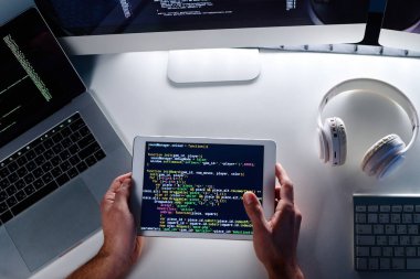 Bilgisayarlı, kulaklıklı ve klavyeli masada otururken ekranda şifreli veri bulunan tableti tutan erkek bilişim mühendisinin elleri yukarıda.