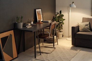 Duvarın yanında duran koyu gri renkli orta boy masa. Oturma odasında konforlu kanepe ve yerli bitkilerle soyut resimler var.