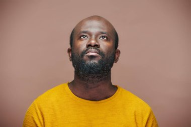 Afro-Amerikalı bir adamın portresi kahverengi arka planda durup düşünceli bir ifadeyle yukarı bakıyor.
