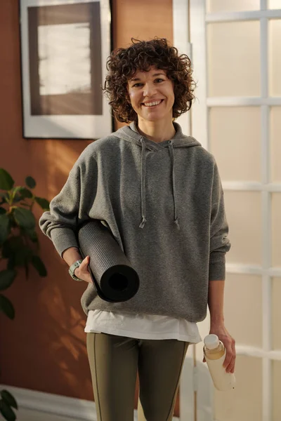 身穿灰色运动服的快乐年轻而活泼的女人 站在客厅里靠着墙和门 看着镜头 手里拿着滚动的垫子 — 图库照片