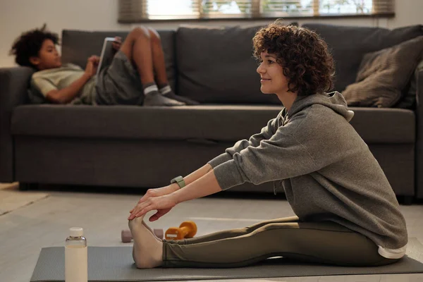 身穿活动服的年轻快乐的黑发女人坐在客厅地板上的垫子上 把胳膊和腿向前伸着 靠着她的儿子 — 图库照片