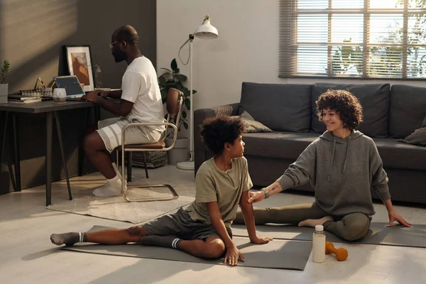 快乐的年轻妈妈穿着活动的衣服坐在客厅地板上的垫子上 和她的儿子在运动过程中交谈 而不是和社交圈的男人在一起 — 图库照片