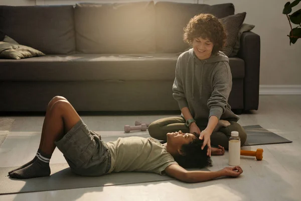 快乐的年轻妈妈穿着灰色的运动服 抚摸着她儿子的头发 晨练过后在客厅地板上的垫子上放松 — 图库照片