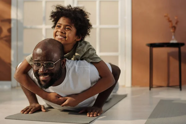 早上在客厅地板上锻炼的时候 可爱而快乐的非裔美国男孩抱着他的父亲在垫子上做俯卧撑 — 图库照片