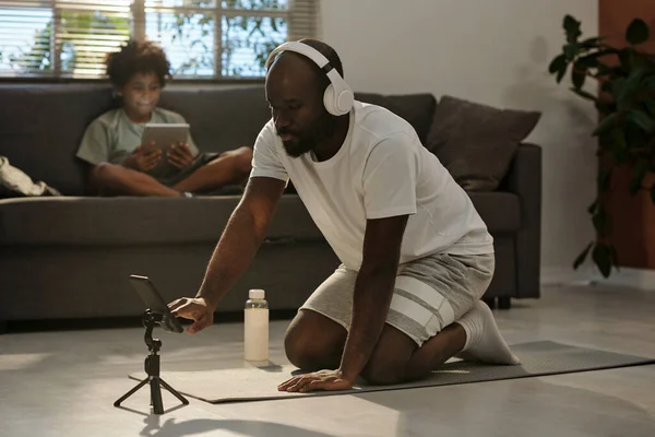 戴耳机的年轻人跪在客厅的垫子上 搜寻健身的网上视频 — 图库照片