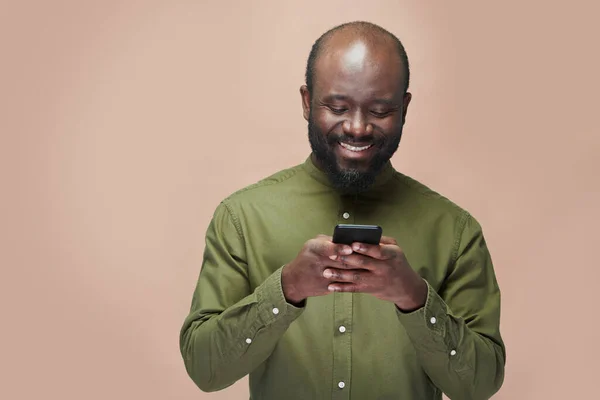 身穿绿色衬衫的非裔美国人在智能手机上读着短信 笑着站在褐色背景上 — 图库照片