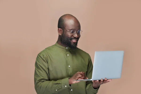 微笑着拿着笔记本电脑在键盘上打字的非裔美国人 站在褐色的背景上 — 图库照片