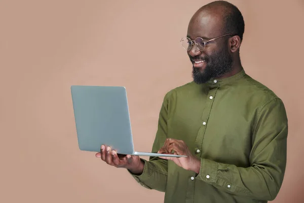 アフリカ系アメリカ人の男性が眼鏡でオンラインで働いている彼のラップトップを使用して笑顔茶色の背景に立って — ストック写真