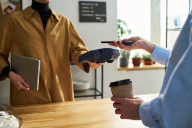 Tezgahı tezgahın üstünde olan tezgahtara yakın çekim ve kafede kahve için yapılan kesintisiz ödeme sırasında akıllı telefonlu kadın müşteri.