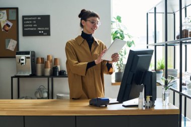 Kafenin genç katibi bilgisayarın başında beklerken tablet kullanıyor ve online siparişler alıyor ya da maliyetleri sayıyor.