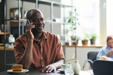 Akıllı telefonlu mutlu Afro-Amerikalı bir adam rahat bir kafede oturmuş, kek ile çay içerken birileriyle konuşuyor.