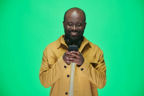 アフリカ系アメリカ人の男性が携帯電話でメッセージを読み 緑の背景に笑顔で立っている — ストック写真