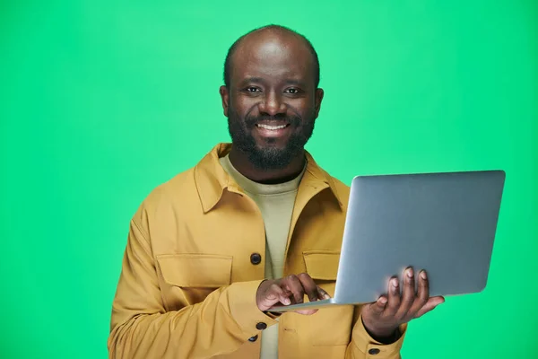 穿着黄色夹克的非裔美国人在绿色背景下使用笔记本电脑时对着相机微笑的肖像 — 图库照片