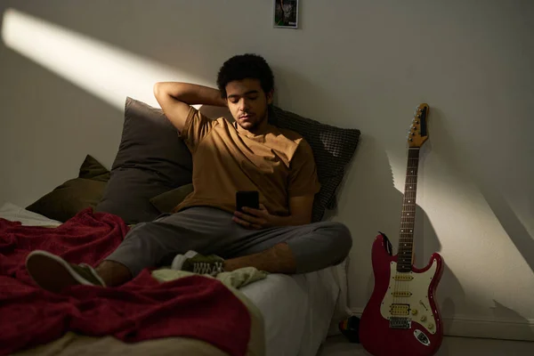 忧心忡忡的年轻人坐在床上 用智能手机看视频 — 图库照片
