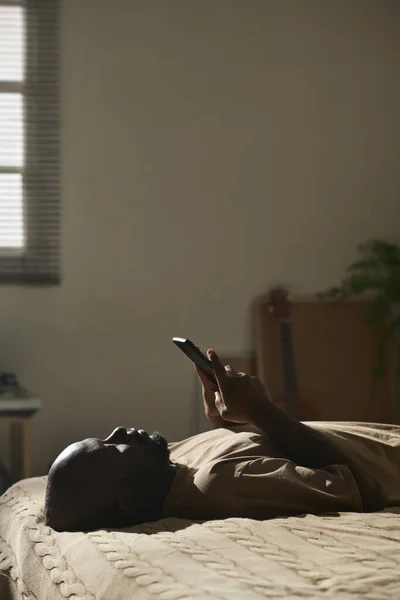非洲裔美国人忧郁男子躺在卧室躺在床上用智能手机阅读信息的垂直图像 — 图库照片