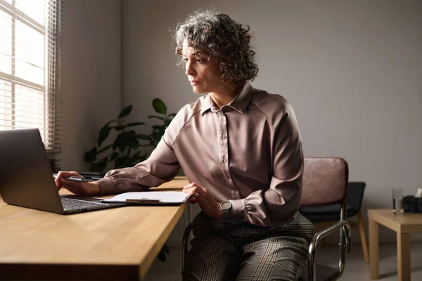 患者の個人データを見てメモを取りながらノートパソコンの前で職場に座っているエレガントな成熟した女性心理学者 — ストック写真