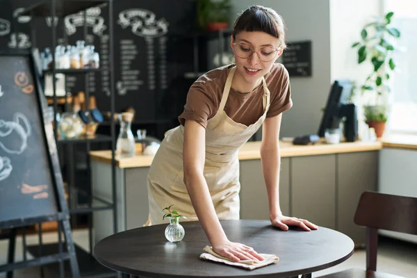 围裙上年轻微笑的女服务员弯腰在小圆桌上擦拭着 同时为咖啡店的新客人们做准备 — 图库照片