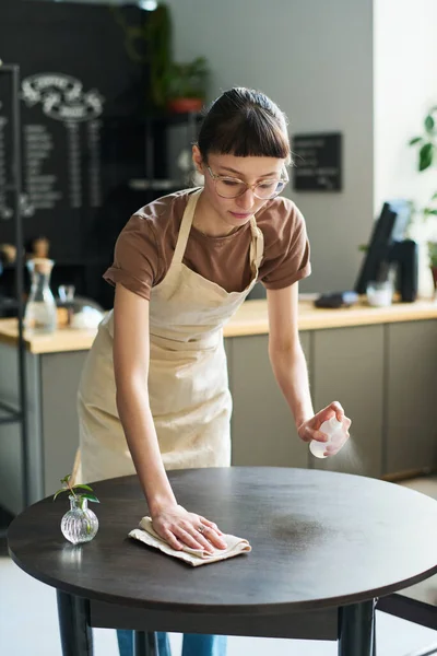 Young brunette woman in apron spraying sanitizer on surface of table and cleaning it with wipe while preparing place for new clients of cafe