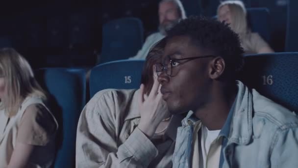年轻的黑人男子和他的白人女友在电影院一起看电影 在彼此耳边低声谈论着这一幕 — 图库视频影像